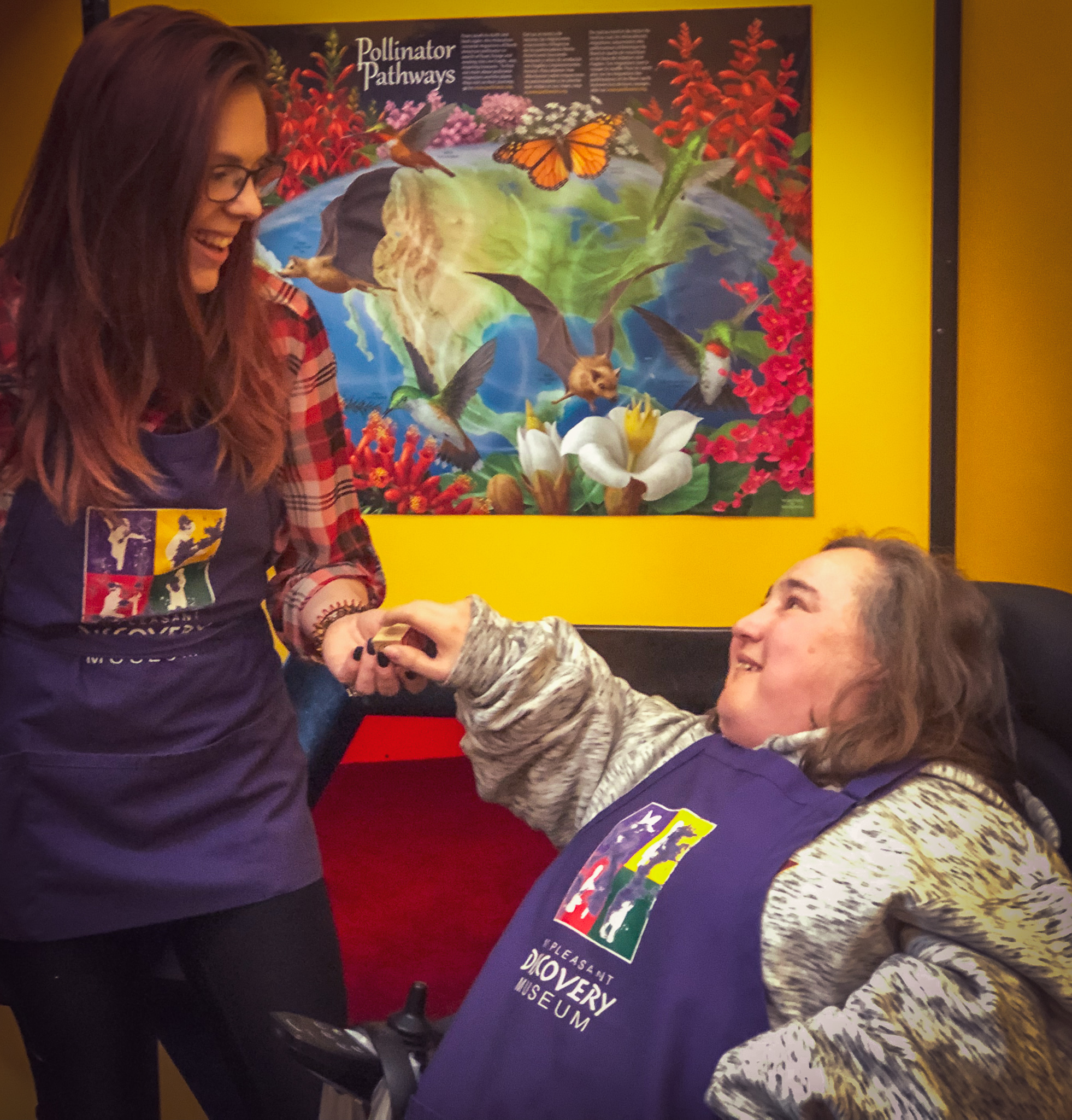 Woman smiling with red hair wearing purple apron working with a woman smiling with brown hair in a purple apron who uses a wheelchair.